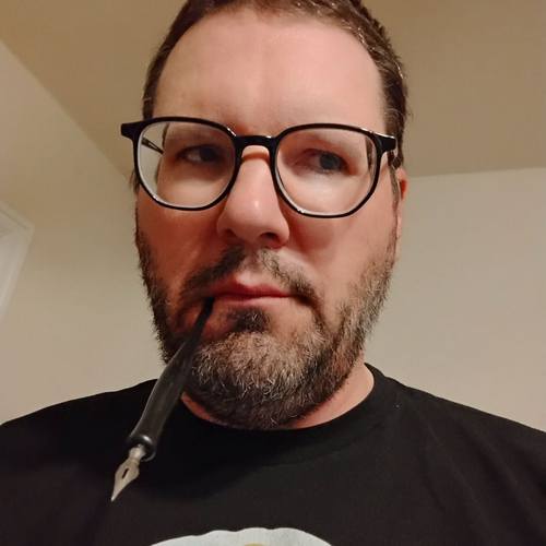 The face of a man with glasses and short, dark, scruffy beard and hair holding in his mouth a dip pen with a bowl-shaped nib and looking off to the side. 