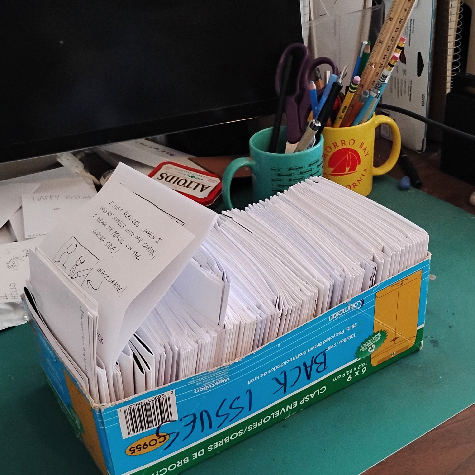 Lots of mini-comics in a tattered box with Back Issues scrawled on it in marker. In the background, a cluttered desk, an Altoids box, artists tools in coffee cups. One cup says Morro Bay California. One says Bianchi. 