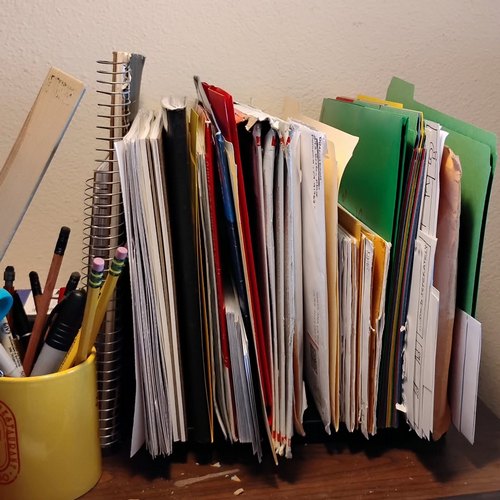 Spiral notebooks, papers, a comp book, envelopes, folders in a vertical organizer. Pens, pencils, a scissors and a ruler in a coffee mug. 