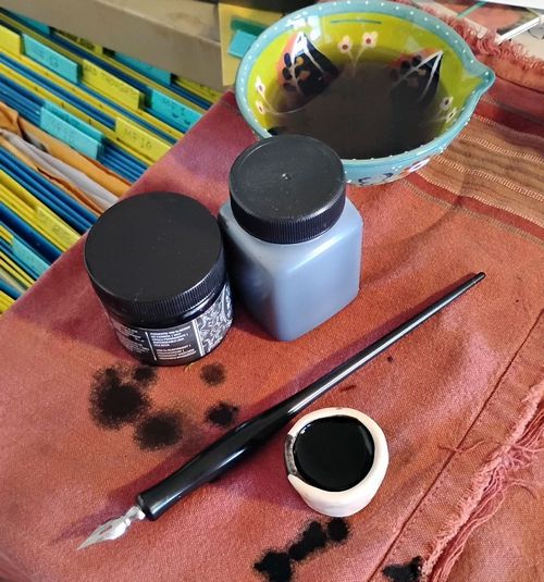 A dip pen, a small pot filled with ink, 2 bottles of ink, and a dish of black water, all laid out on an old dishtowel stained with drops of black ink.